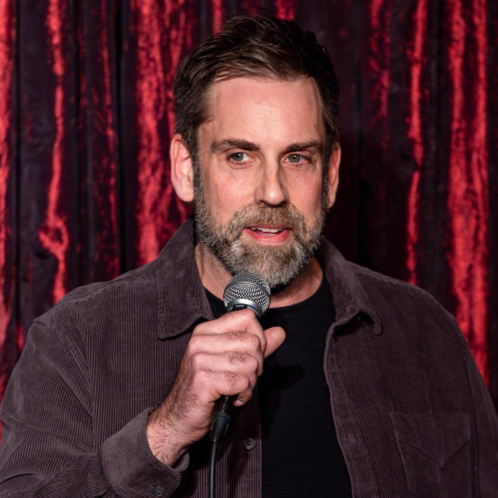 SIB Comedy performing stand-up onstage in front of red curtains